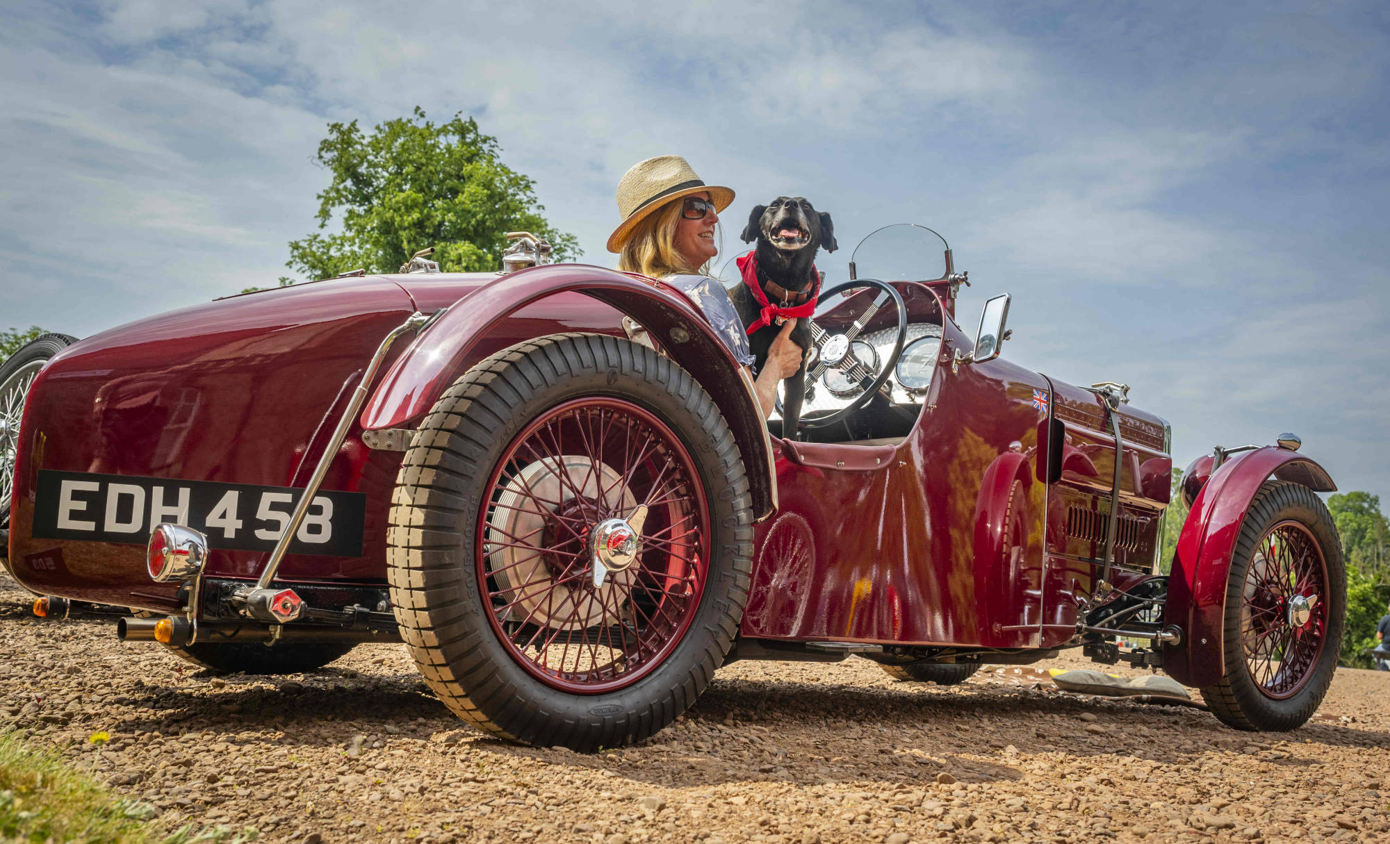 Ruby, a deep red MG TA Special with cycle wings and a tapered tail, parked on grass at a classic car rally, a passenger and a small dog in the cockpit.